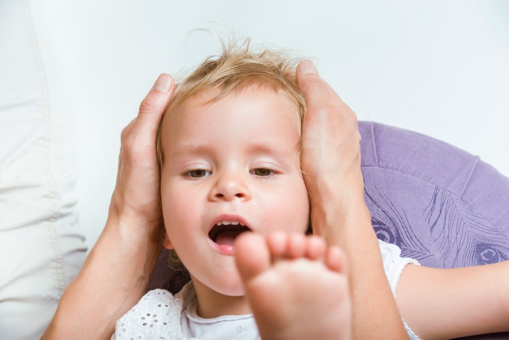 Therapeutin Miriam Wagner behandelt den Kopf eines zweijährigen Mädchens mit beiden Händen an den Schläfen welches auf ihrem Schoß liegt und zu ihr herauf schaut. Ihre linke Fußsohle erscheint im Bild